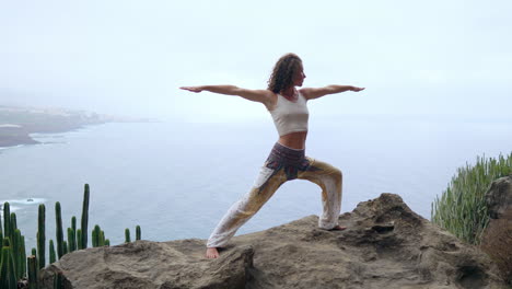 Engaged-in-the-yoga-warrior-pose-by-the-ocean,-beach,-and-rocky-mountains,-the-woman-epitomizes-motivation,-inspiration,-and-a-health-conscious-lifestyle